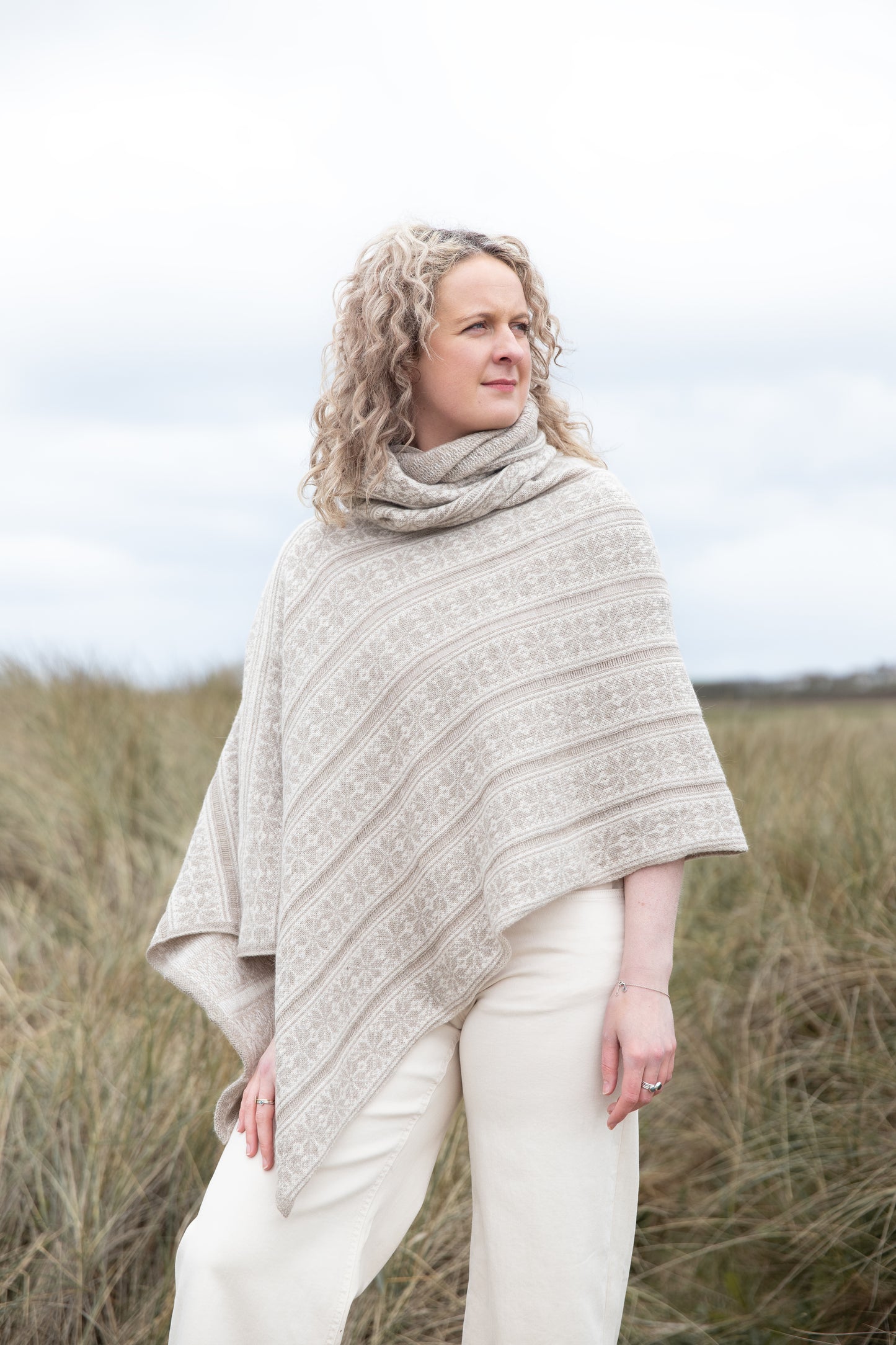 
                  
                    Woman wearing a beige all over fairisle pattern ponchi on some dunes
                  
                