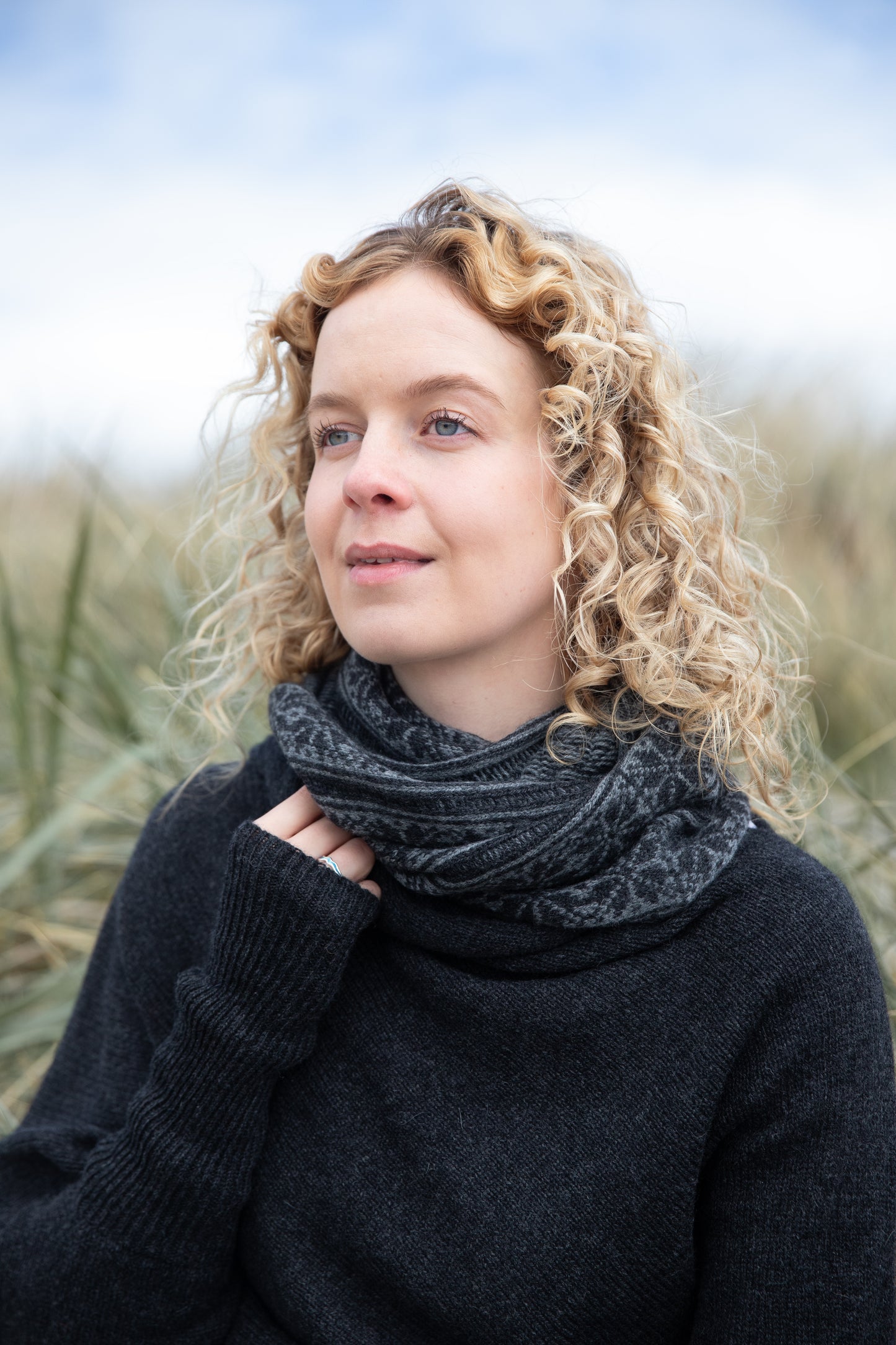 
                  
                    Woman wearing all over fair isle snood in charcoal and grey colour way on an Orkney beach
                  
                