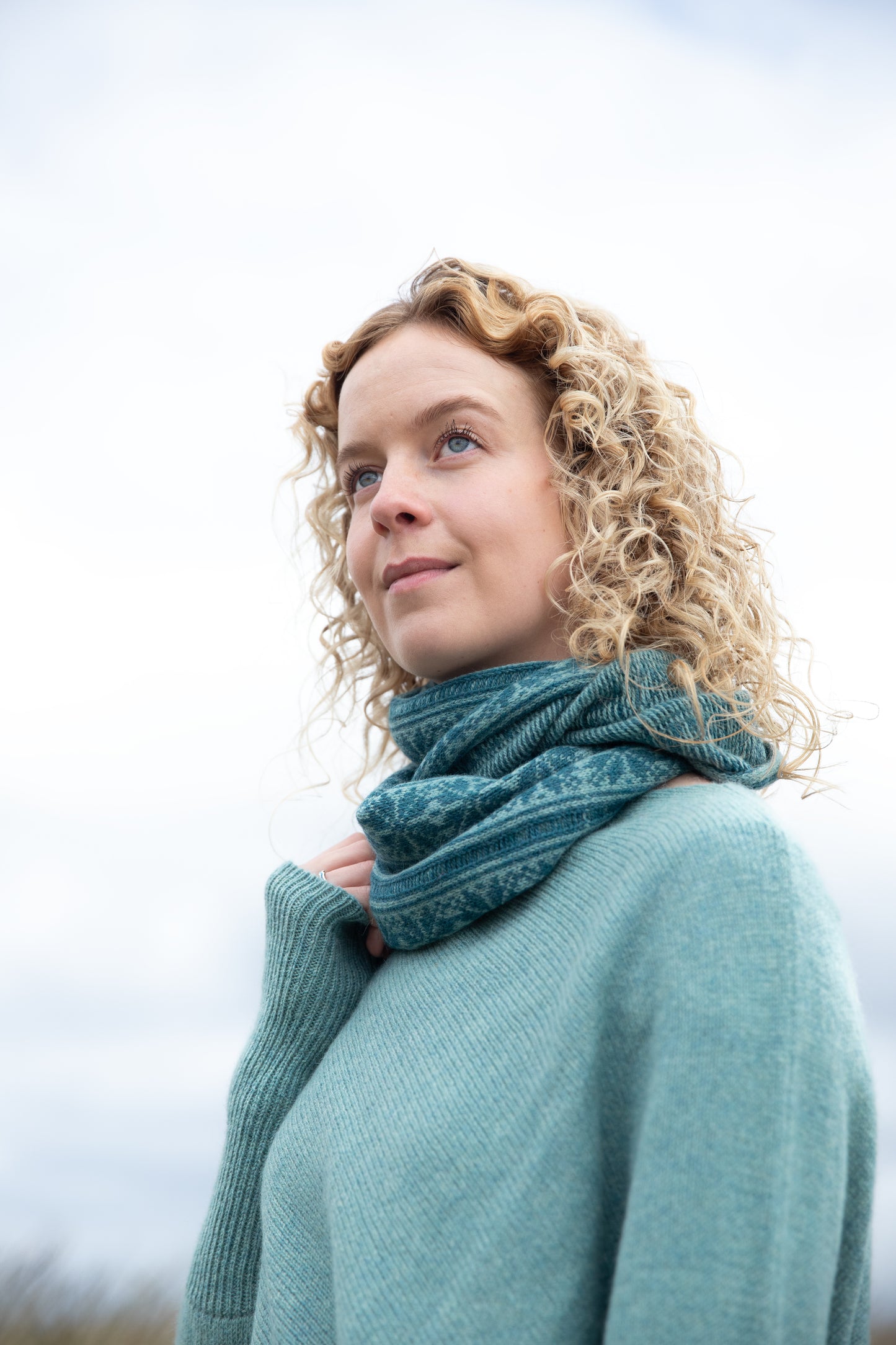 
                  
                    Woman wearing all over fair isle snood in Basil and Shrub colour way on an Orkney beach.
                  
                